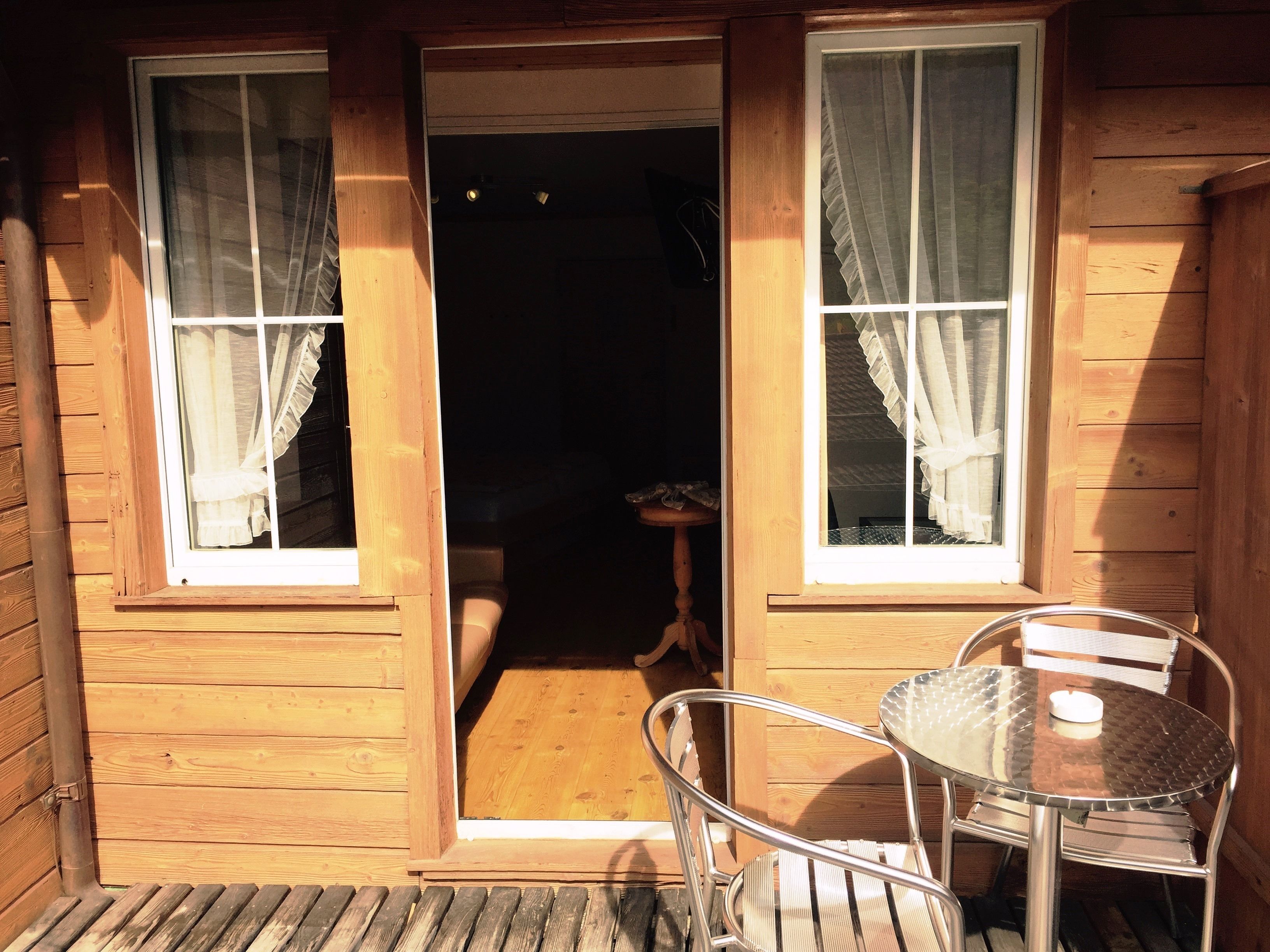 A wooden cabin with a glass door leading to a small room. There is a metal table and a chair outside the door.