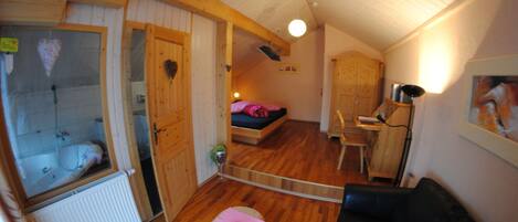 Une pièce chaleureuse avec un plafond en bois, un lit recouvert d’une couverture rose, un canapé en cuir noir et une petite table sur laquelle se trouve un objet rouge.