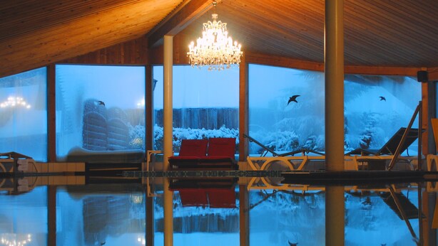 Piscina interna com teto de madeira, um candelabro e vista para uma paisagem nevada através de grandes janelas.
