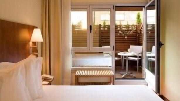 Quarto de hotel com cama, mesa de cabeceira, luminária, um banco e uma porta de vidro deslizante que dá para uma área de estar externa.