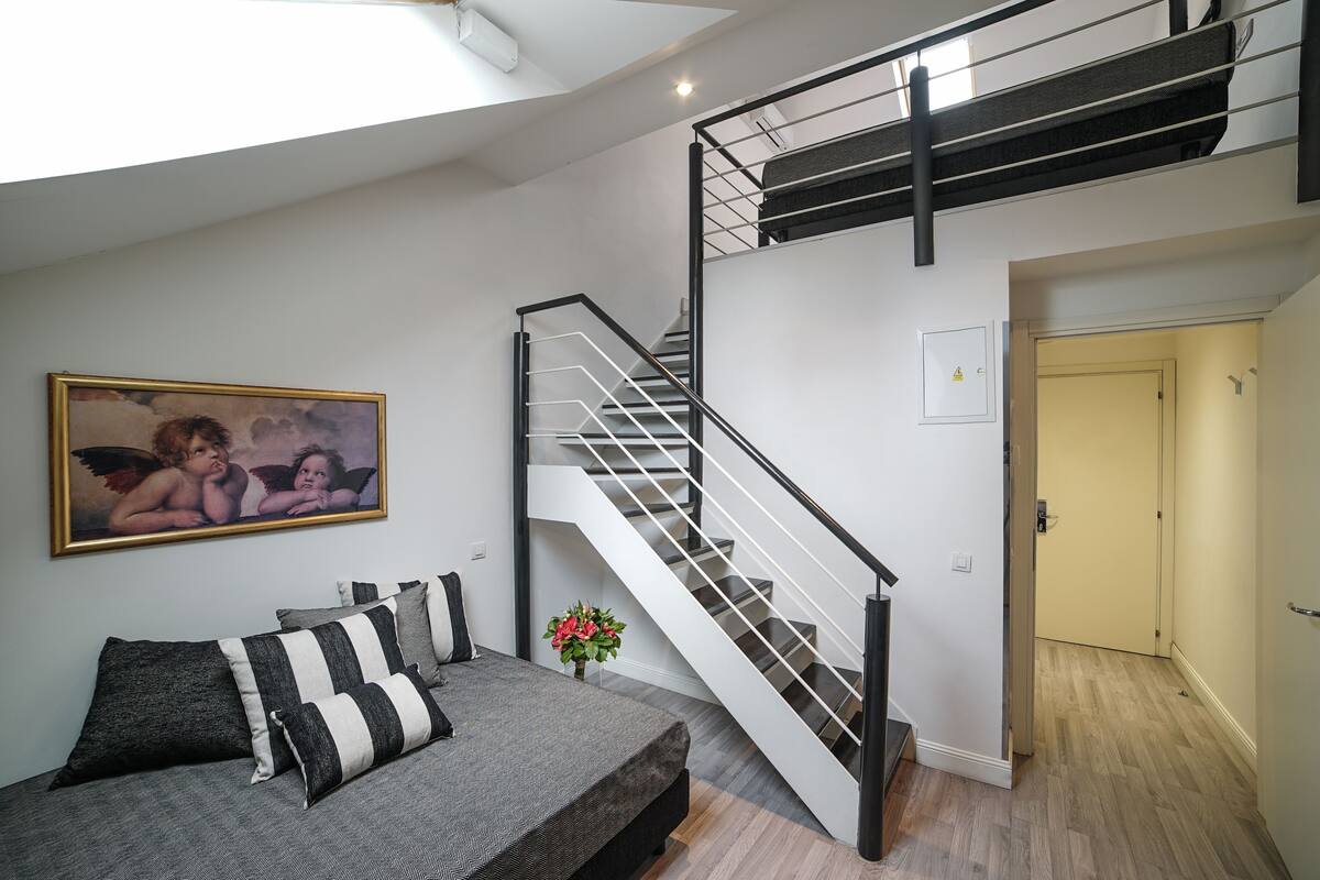 A modern bedroom with a grey sofa, black and white striped pillows, a painting of cherubs, and a staircase with a black railing.