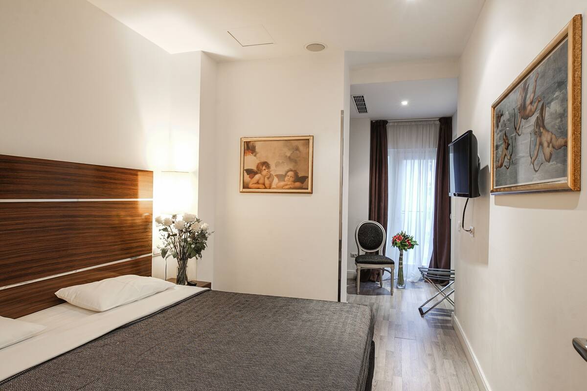A hotel room with a wooden headboard, a bed with a patterned bedspread, a painting on the wall, a television, and a chair with a vase of flowers.