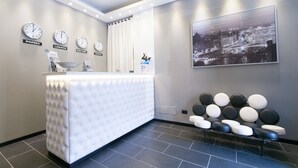 A reception area with a white reception desk, clocks indicating different cities, a large framed cityscape, and a row of black and white chairs.