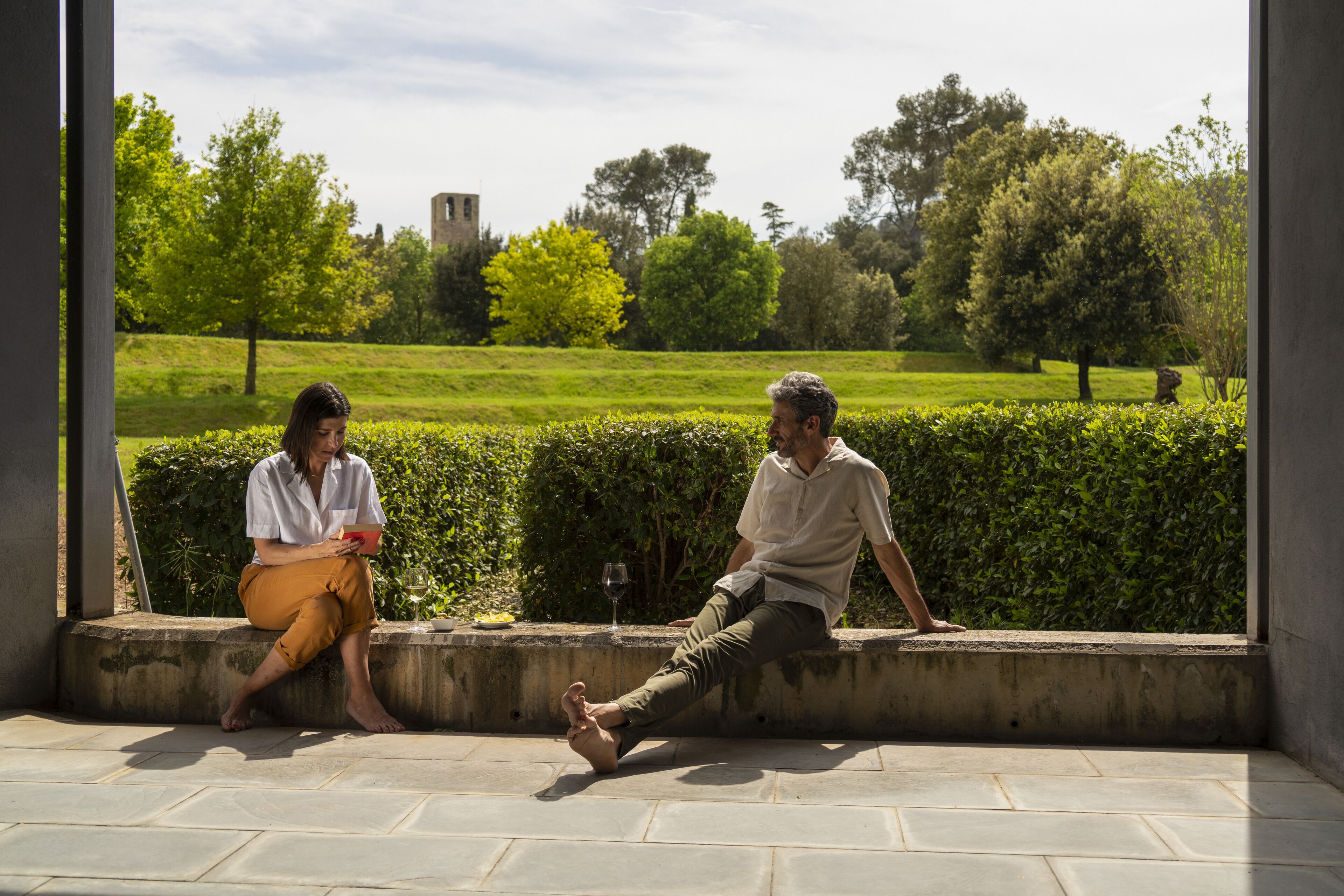 Two people sitting on a concrete ledge with a view of a green park and a distant building.
