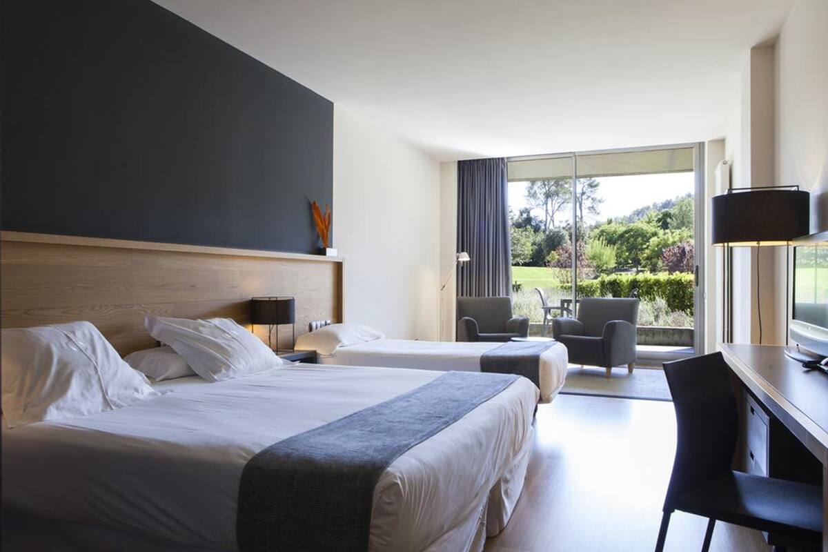 Habitación de hotel con dos camas, un escritorio y una silla. Se observa un amplio ventanal con vistas al follaje exterior.