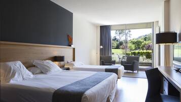 A hotel room with two beds, a desk, and a chair. There is a large window with a view of greenery outside.