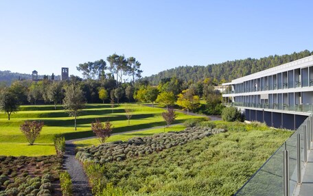 A modern building with a glass facade, surrounded by well-maintained gardens and a clear view of a forested hill in the background.