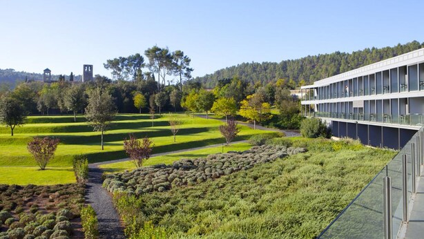 Un edificio moderno con fachada de vidrio, rodeado de jardines cuidados y con vista despejada a una colina boscosa al fondo.