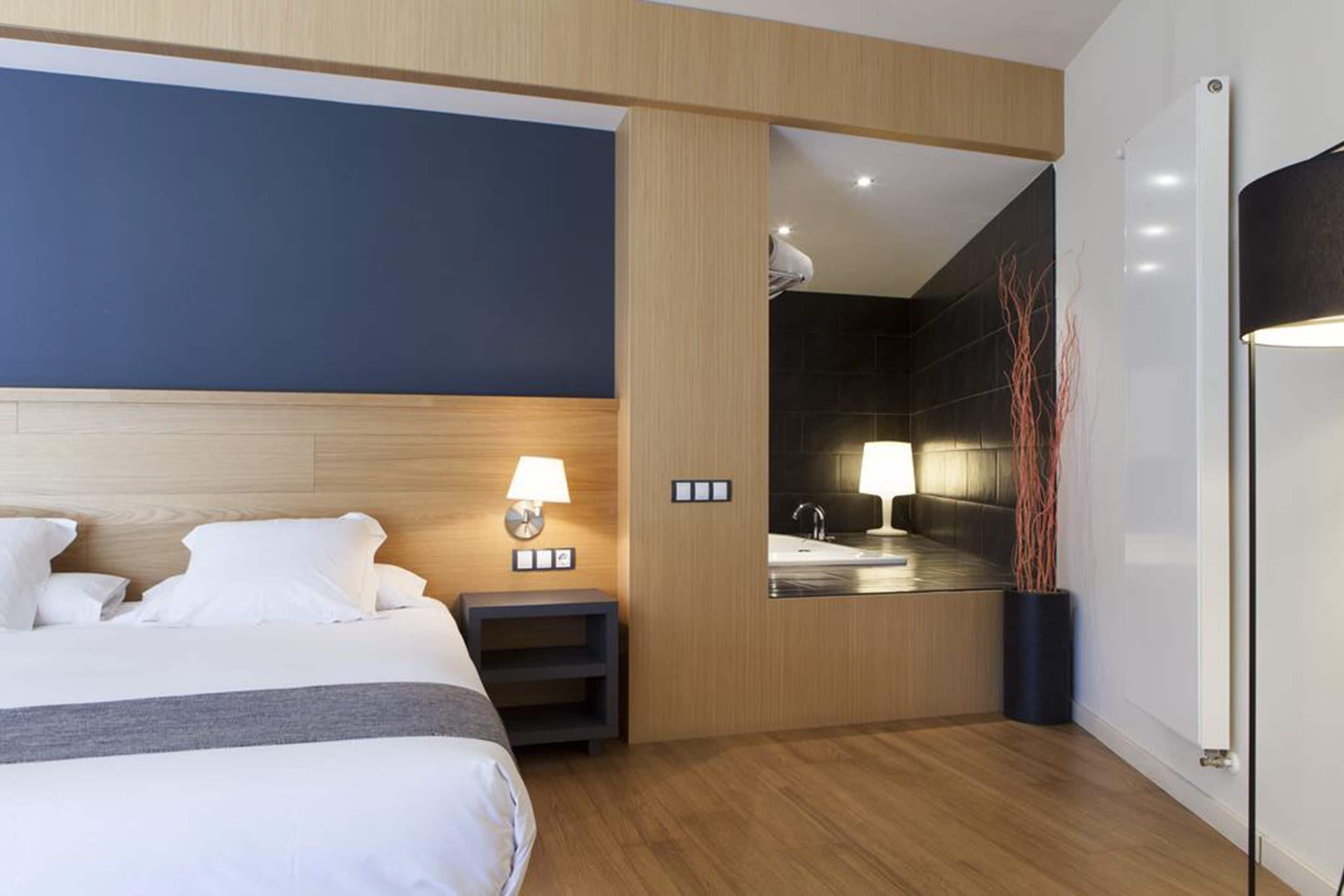 A modern hotel room with a large bed, bedside table, lamp, and a view of the bathroom.