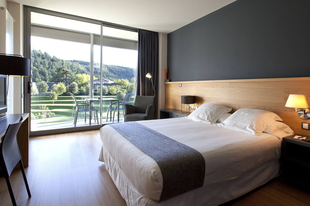Habitación de hotel con una cama grande, un escritorio con una silla, una mesita de noche con una lámpara y un teléfono. Se observa un paisaje verde a través de la ventana.