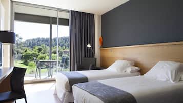 Habitación de hotel con dos camas, un escritorio y una silla. Amplia ventana con vistas a zonas verdes y un balcón.