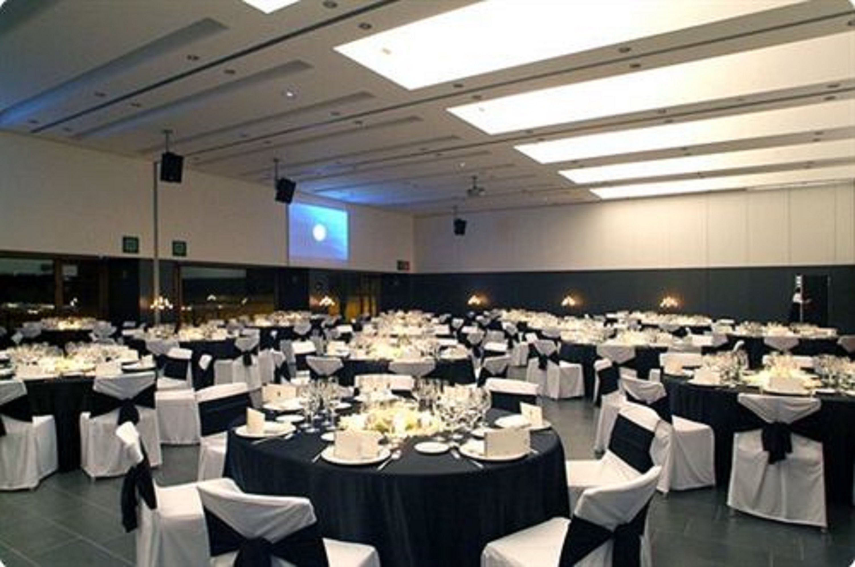 Un amplio salón de banquetes con mesas redondas dispuestas para comer, sillas blancas y manteles negros.
