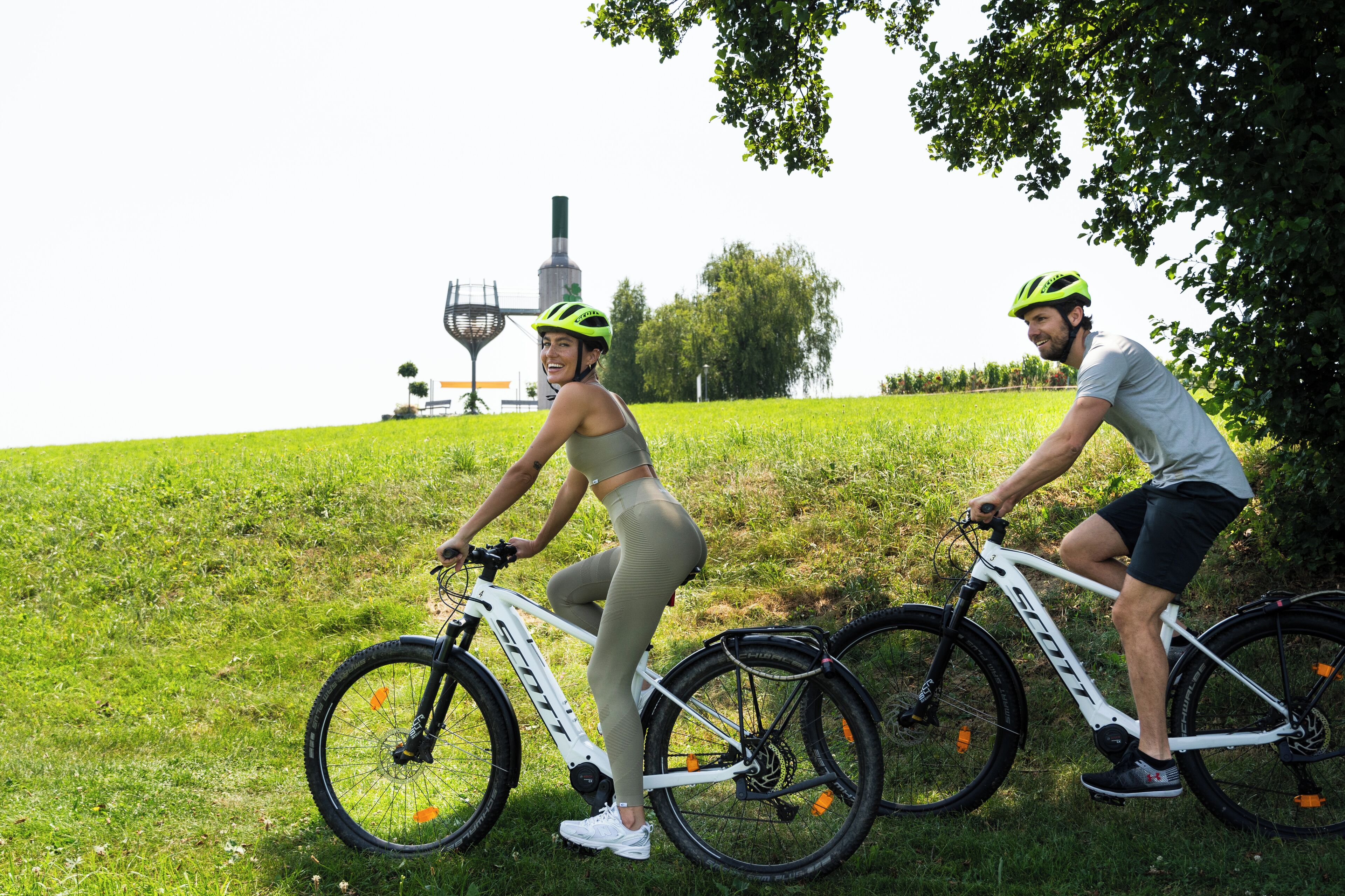 Dos personas en bicicleta en una zona cubierta de hierba, con una estructura al fondo.