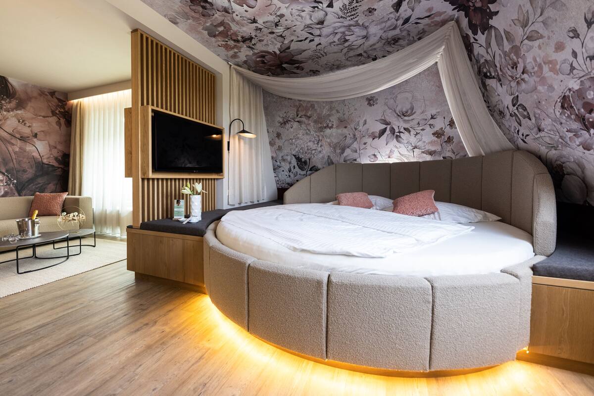 Une chambre moderne avec un lit aux lignes courbes, un téléviseur à écran plat, un mur d’accent en bois et un plafond orné de motifs floraux.