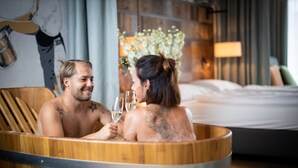 A couple in a wooden bathtub with a bed in the background. - Hotel Vier Jahreszeiten (Loipersdorf bei Fuerstenfeld)