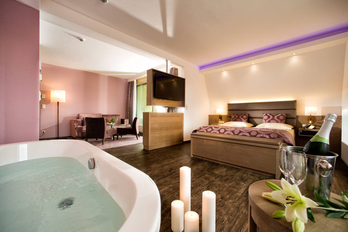 Une chambre d’hôtel moderne dotée d’une grande baignoire, d’un lit avec une tête de lit, d’un bureau et d’un téléviseur fixé au mur.