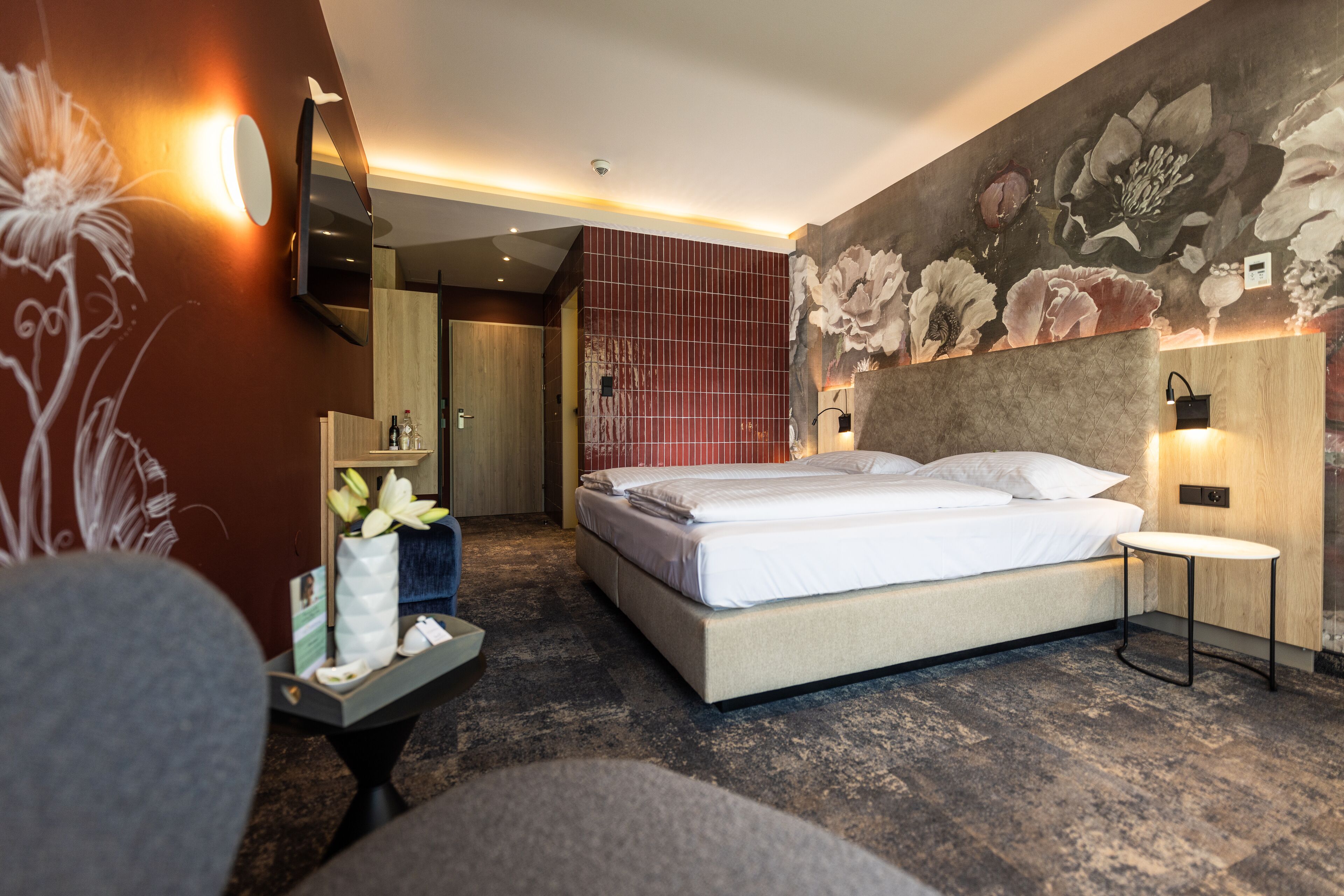 Una habitación de hotel moderna con una cama grande, un televisor de pantalla plana, un cabecero de madera y un mural floral en la pared.