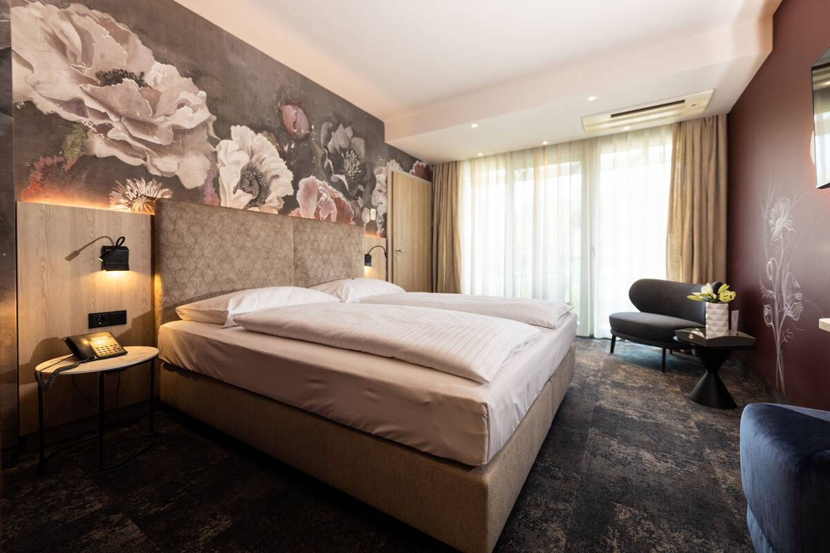 Une chambre d’hôtel avec un grand lit, un téléphone sur la table de chevet, une petite table avec une lampe, une chaise et un mur décoratif orné d’œuvres d’art florales.