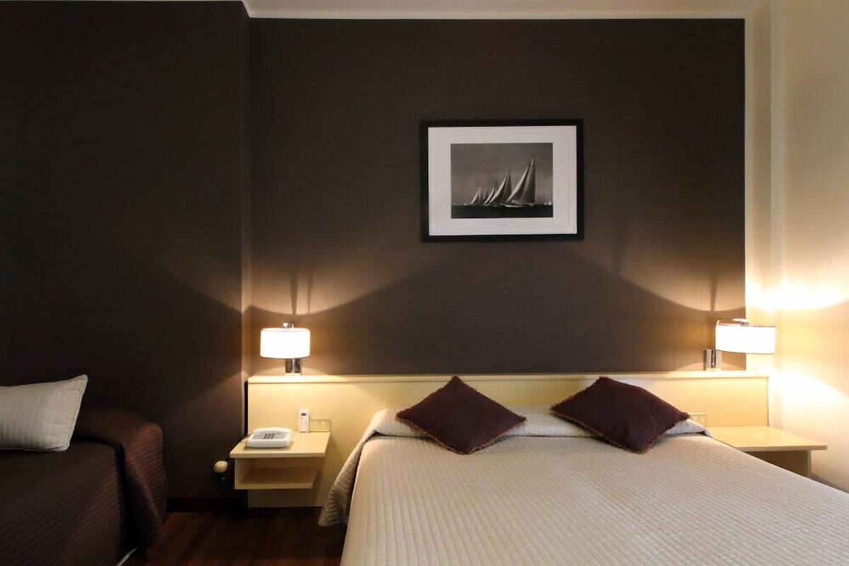 Hotellihuone, jossa on sänky, kaksi yöpöytää, kaksi lamppua, yöpöydän päällä oleva esine ja seinällä oleva kehystetty kuva.