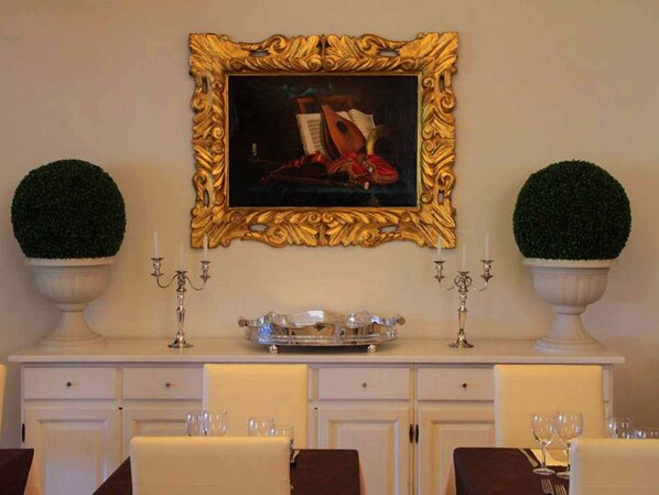 A dining area with a white buffet table, two potted plants, a gold-framed painting, and a candelabra.