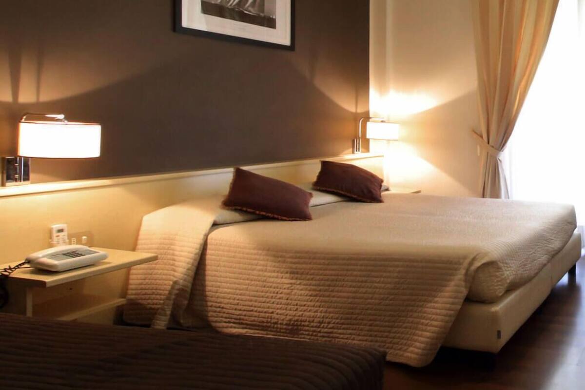 Hotellihuone, jossa on sänky, kaksi yöpöytävalaisinta, yöpöytä, jossa on puhelin, ja seinällä oleva kehystetty kuva.