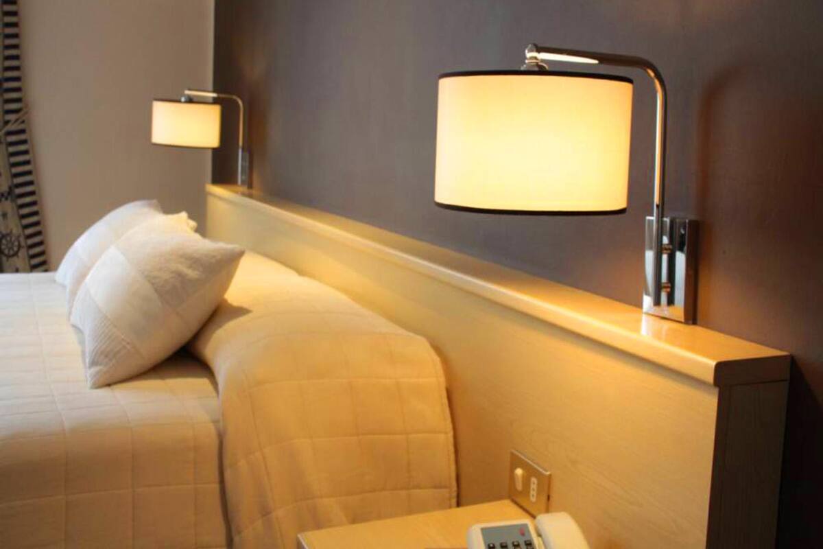 Hotellihuone, jossa on sänky, yöpöytä lampun kanssa ja seinään kiinnitetty valaisin.