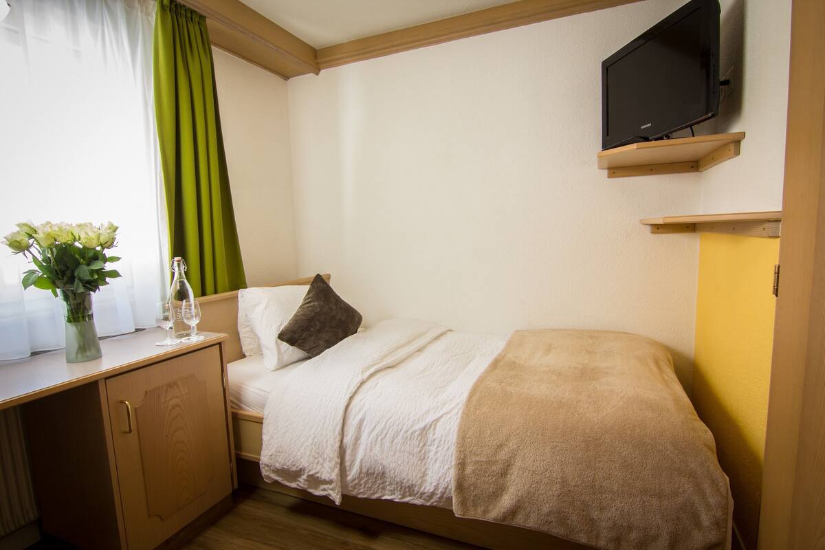 A small, neatly arranged bedroom with a single bed, a bedside table with a vase of flowers, a television mounted on the wall, and a window with curtains.