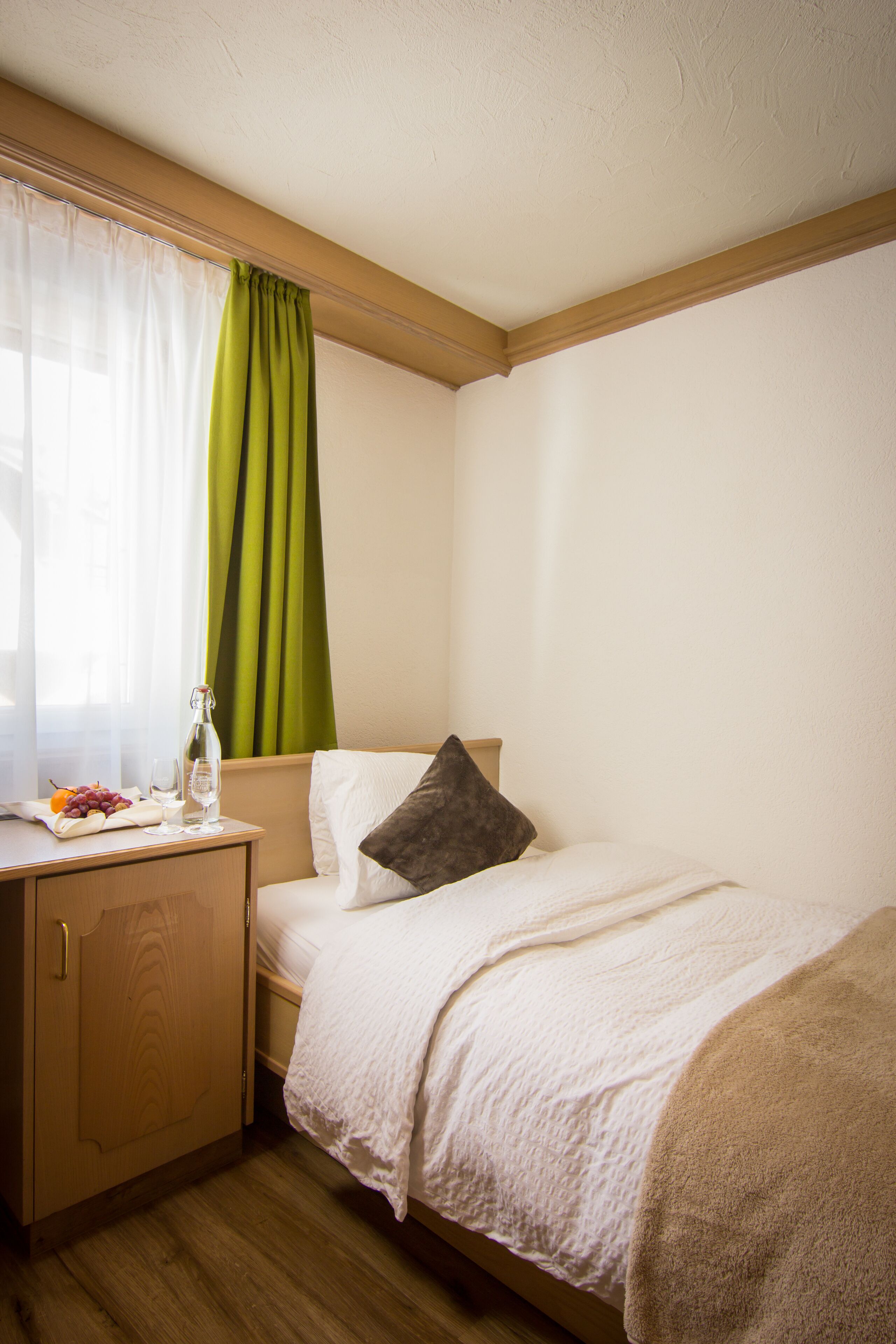 Habitación de hotel ordenada con cama, mesita de noche pequeña con una botella y vasos, cortina verde y ventana con cortinas translúcidas.