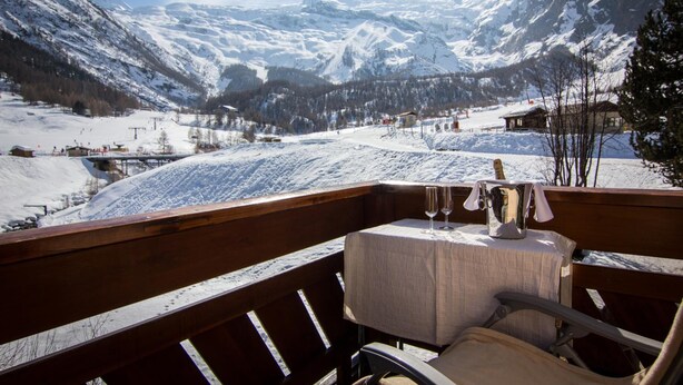 A wooden deck with a table and chairs, overlooking a snowy mountain landscape with ski slopes and buildings. - Hotel Bristol (Saas-Fee)