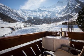 A wooden deck with a table and chairs, overlooking a snowy mountain landscape with ski slopes and buildings. - Hotel Bristol (Saas-Fee)