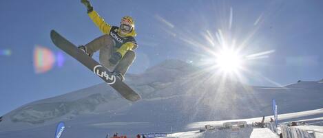 A snowboarder in mid-air performing a jump at a snowy venue with banners and a clear sky.