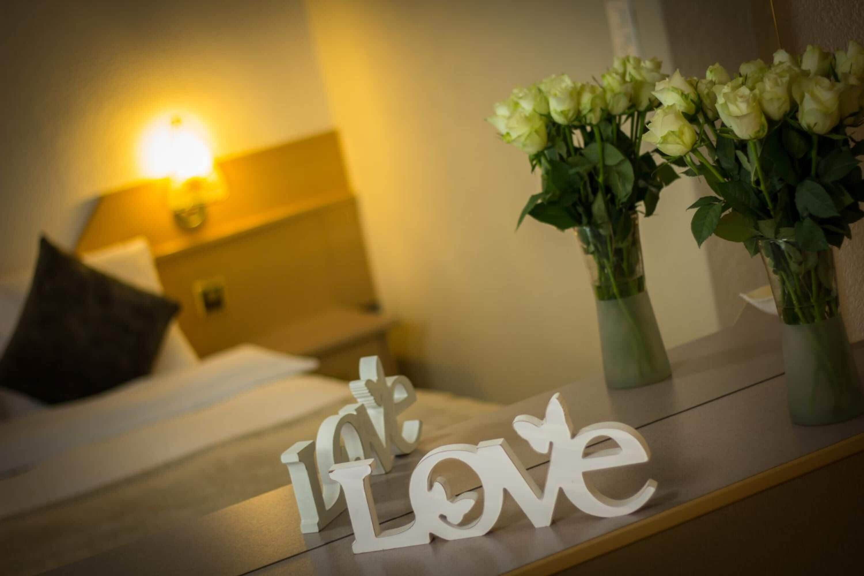 A hotel room with a bed, a nightstand with a lamp, and two vases with white flowers on a nightstand.