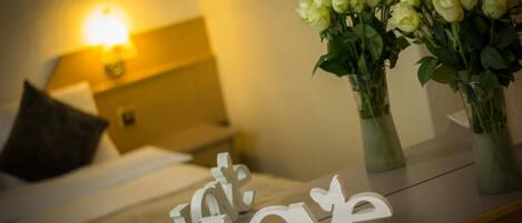 A hotel room with a bed, a nightstand with a lamp, and two vases with white flowers on a nightstand.