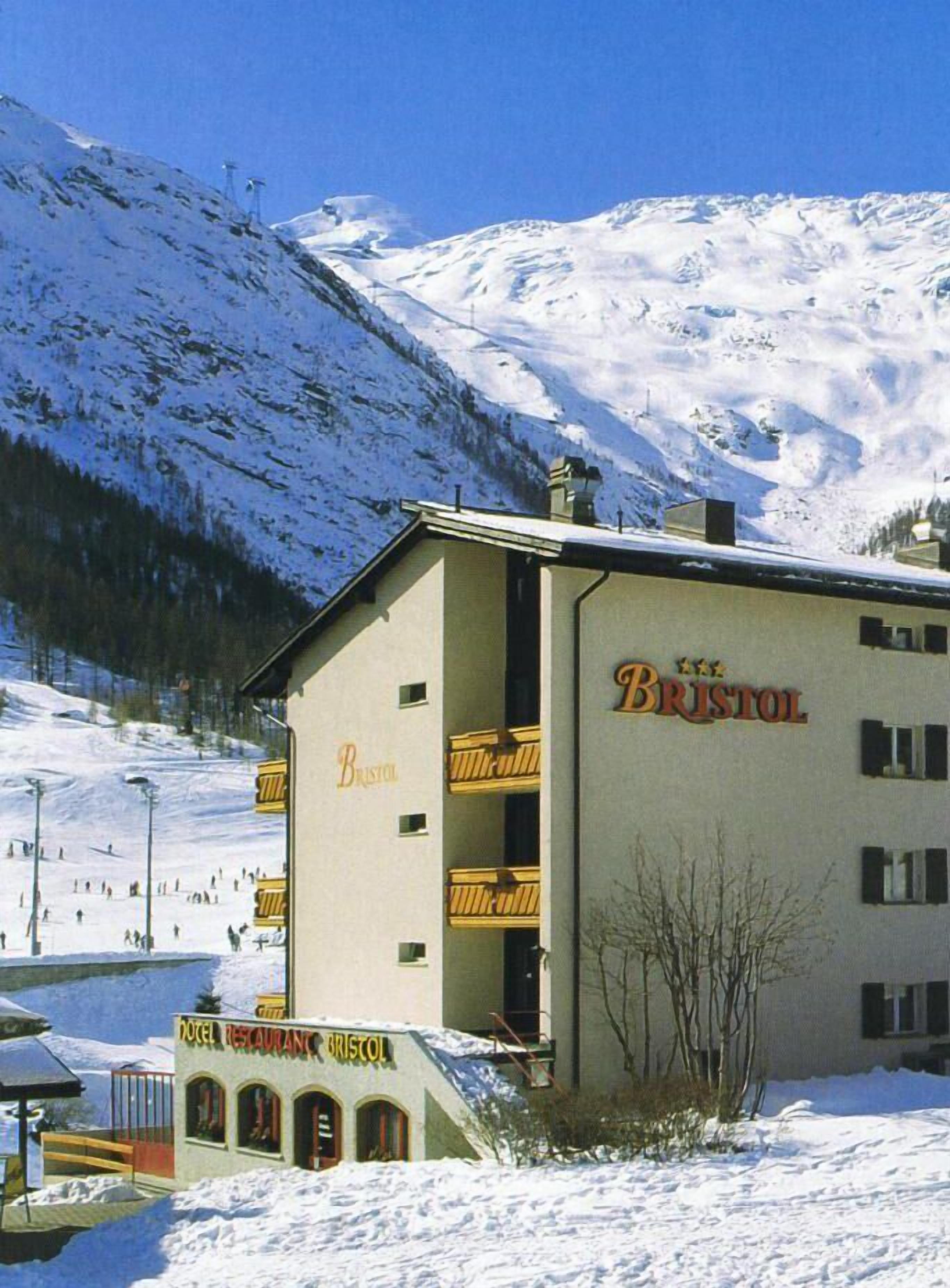 Un bâtiment portant le nom « BRISTOL », avec une enseigne indiquant « HÔTEL-RESTAURANT BRISTOL », et une piste de ski en arrière-plan.