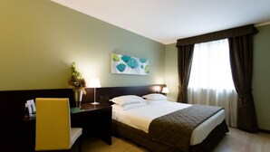 Una habitación de hotel con cama, un escritorio con silla, una mesita de noche, una lámpara, un cuadro en la pared y una ventana con cortinas.
