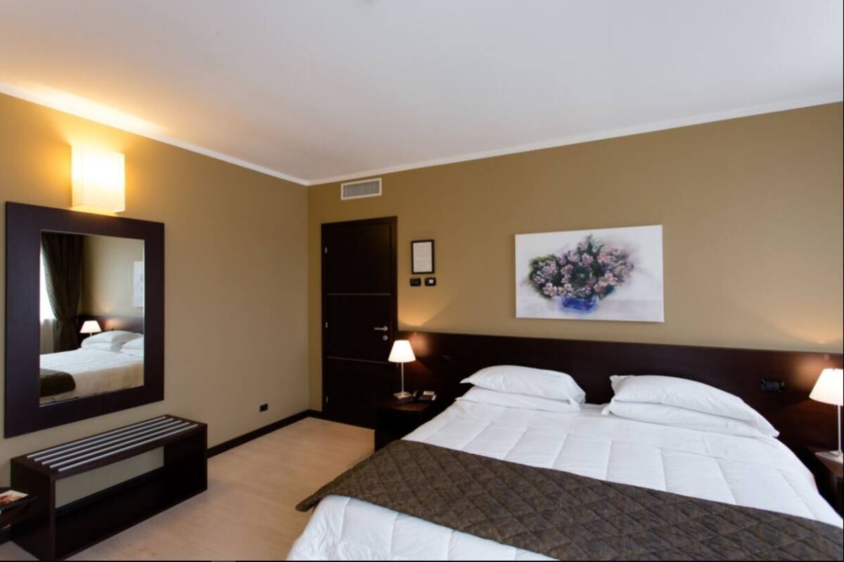 A hotel room with a large bed, a bedside table with a lamp, a bench, a door, and a wall-mounted mirror.