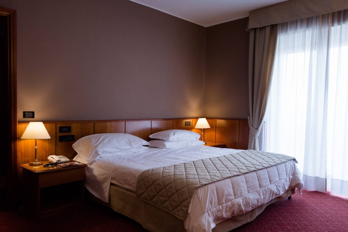 A hotel room with a large bed, two bedside lamps, a nightstand with a phone, and a window with curtains.