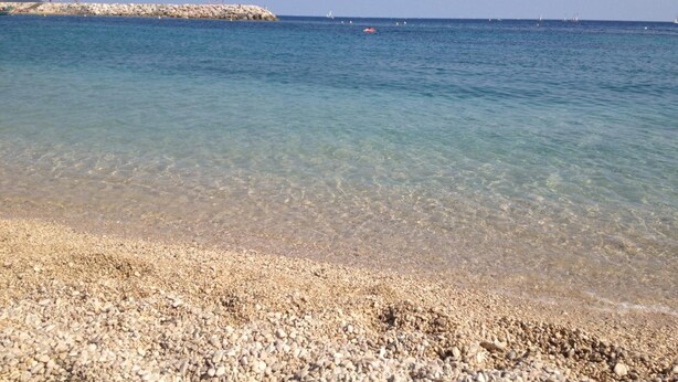 Una playa de guijarros con agua cristalina y un rompeolas rocoso al fondo.