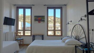 A bedroom with a bed, a chair, a desk, and a TV. There are two windows with a view of the outside.