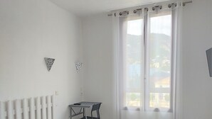 Un dormitorio con cama, una mesita, una silla, un radiador, una ventana con cortinas y un televisor en la pared.