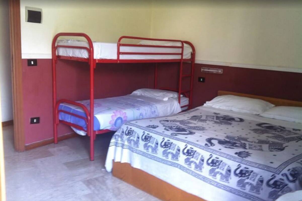 Une chambre avec un lit superposé doté d’une structure en métal rouge, un lit inférieur recouvert d’une couette bleue et blanche, et un lit supérieur recouvert d’une couette blanche.