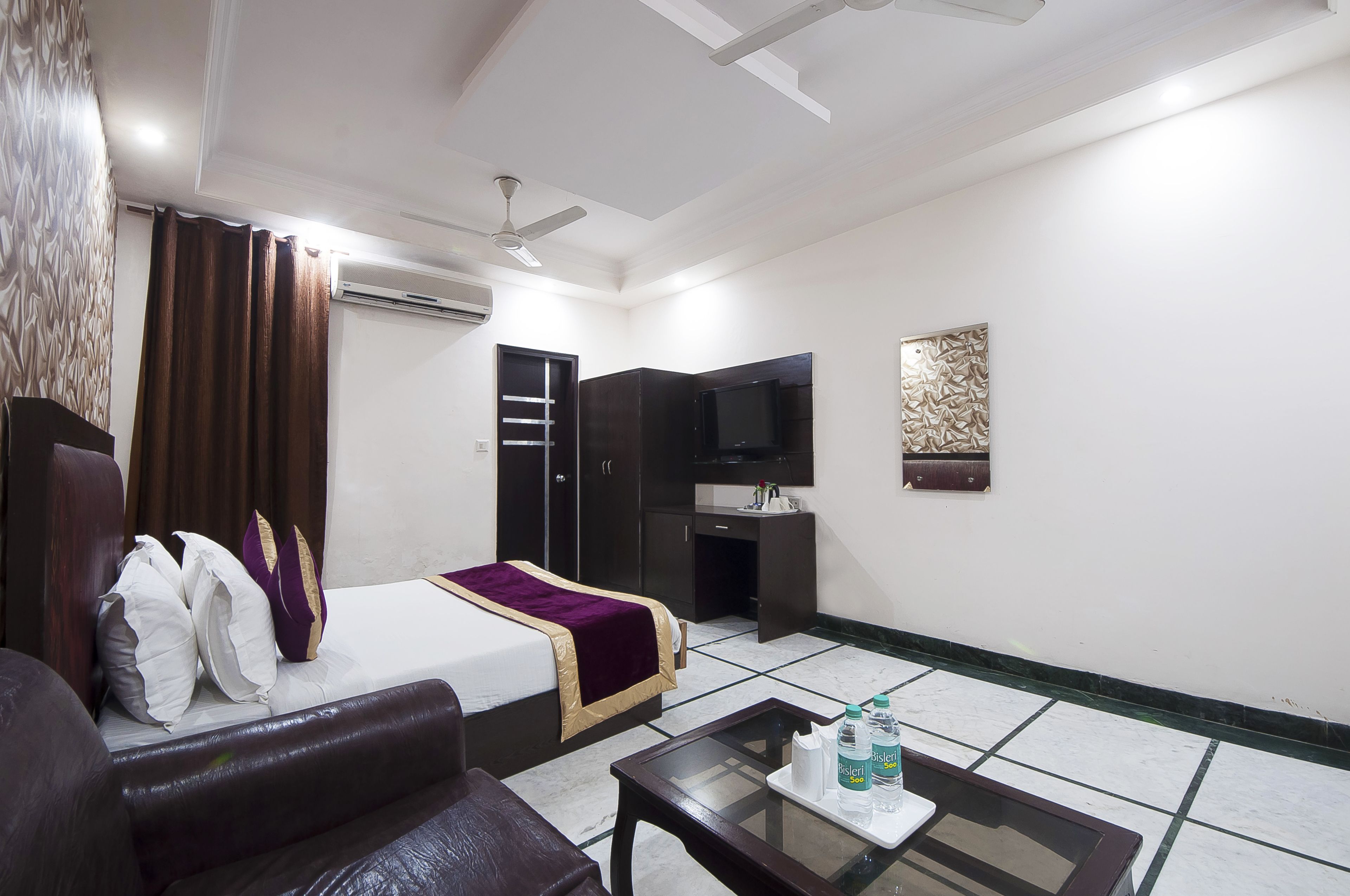 A hotel room with a bed, a sofa, a coffee table with water bottles, a TV, and a ceiling fan.