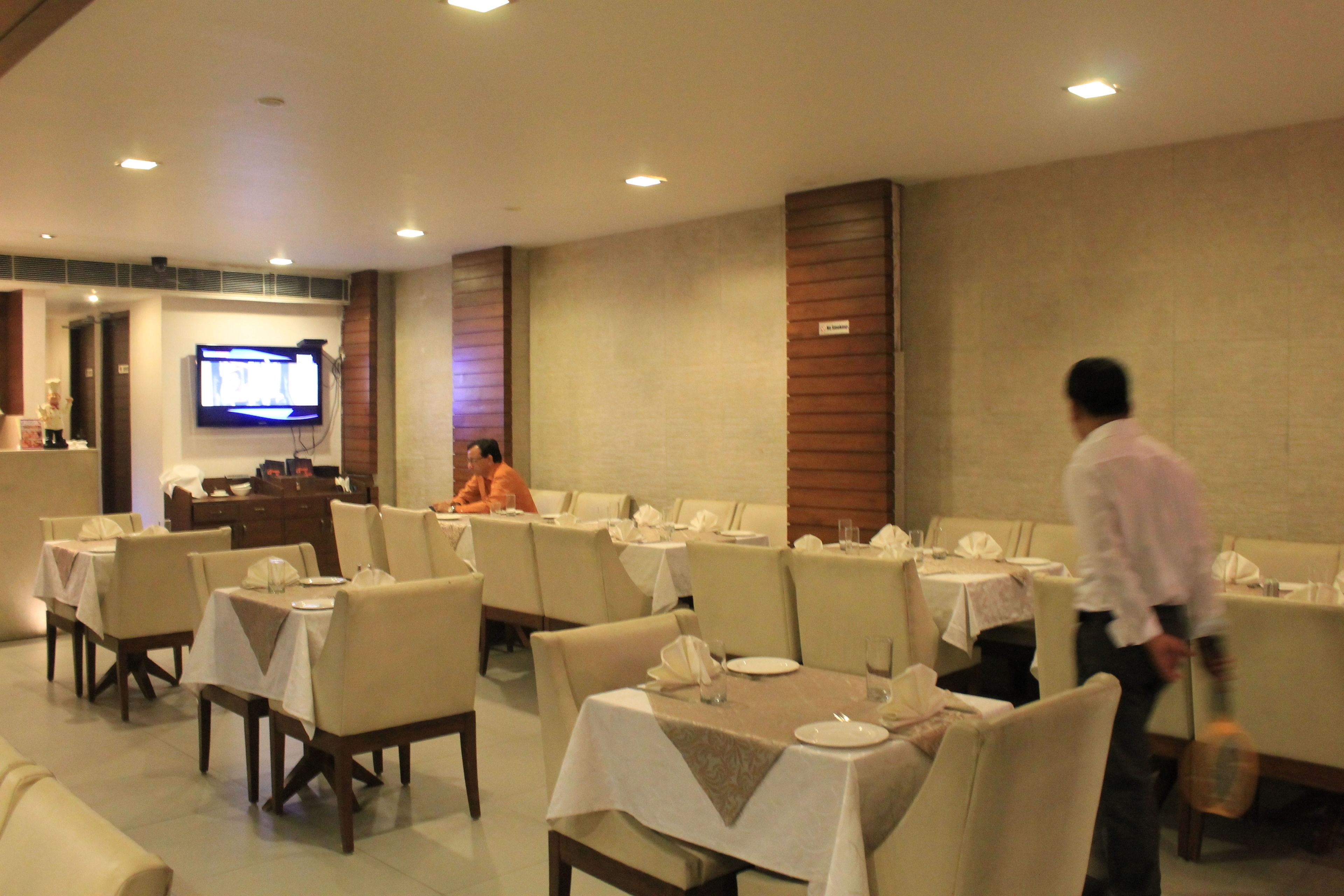 A restaurant with tables set for dining, a television mounted on the wall, and a person walking through the space.