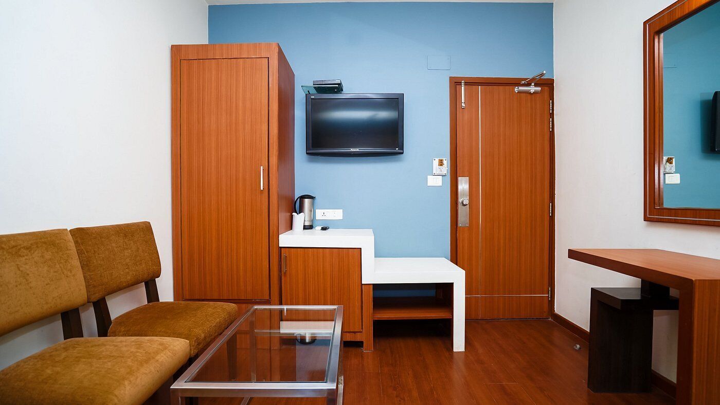 A compact room with a sofa, a small table, a TV mounted on the wall, a wooden wardrobe, and a mirror.