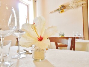 A dining table with a white tablecloth, a glass vase with a white flower, and empty wine glasses. - Hotel VelaSole (Siniscola)