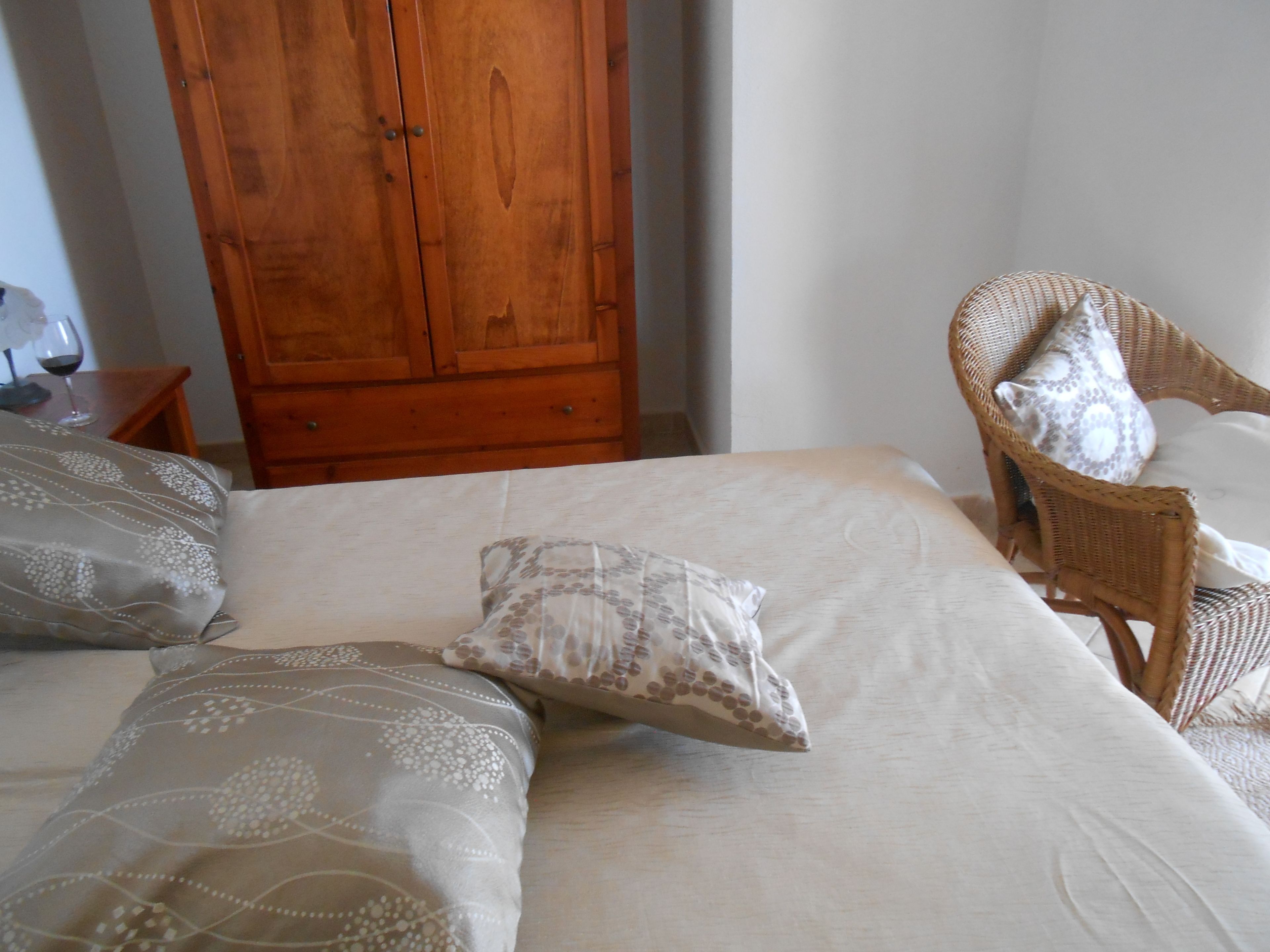 A bedroom with a bed, pillows, a wooden wardrobe, and a wicker chair.