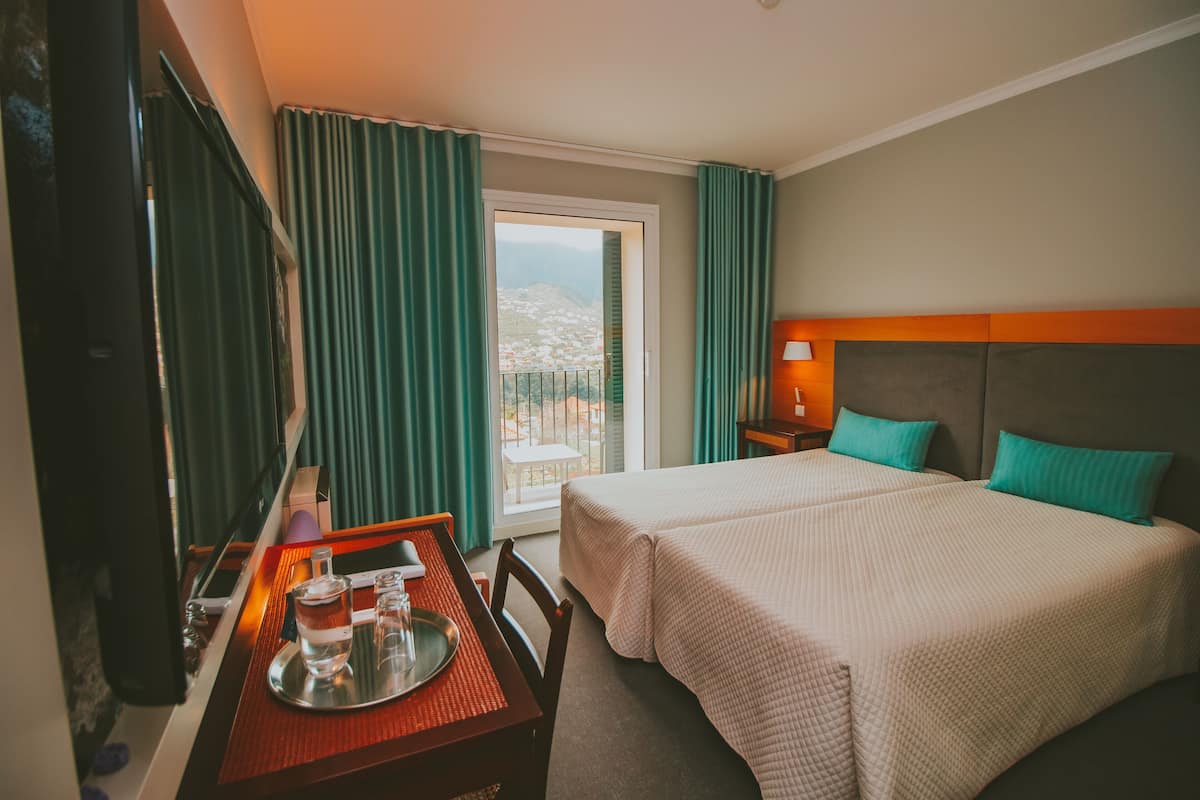 A hotel room with two beds, a desk with a tray of glasses and a bottle, a TV, and a view of a balcony with a cityscape.