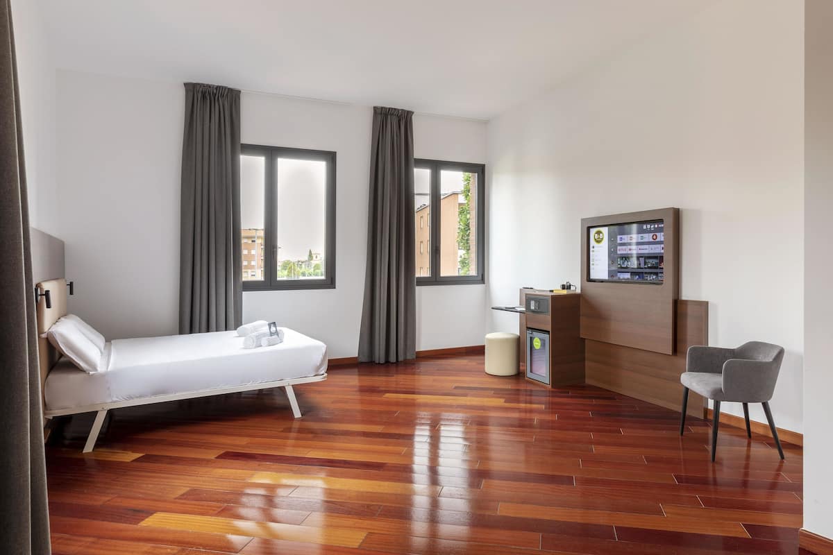 A hotel room with a bed, two windows with curtains, a wooden floor, a desk with a computer, a chair, and a wall-mounted TV.