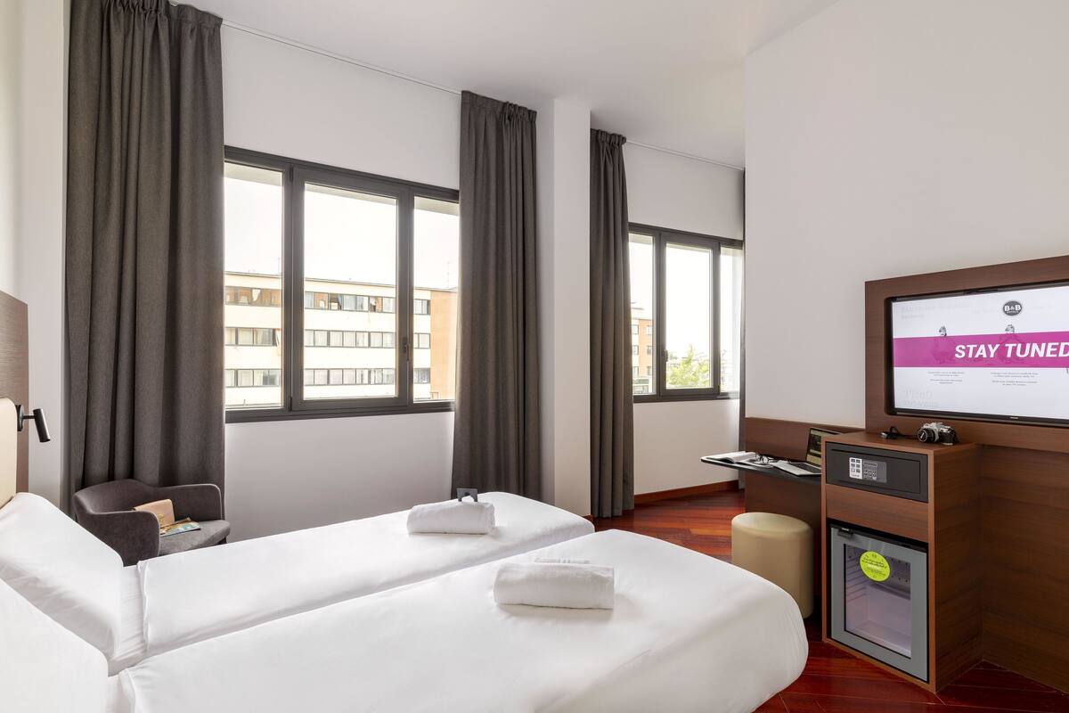 A hotel room with two beds, a desk, a chair, a television, and a minibar.