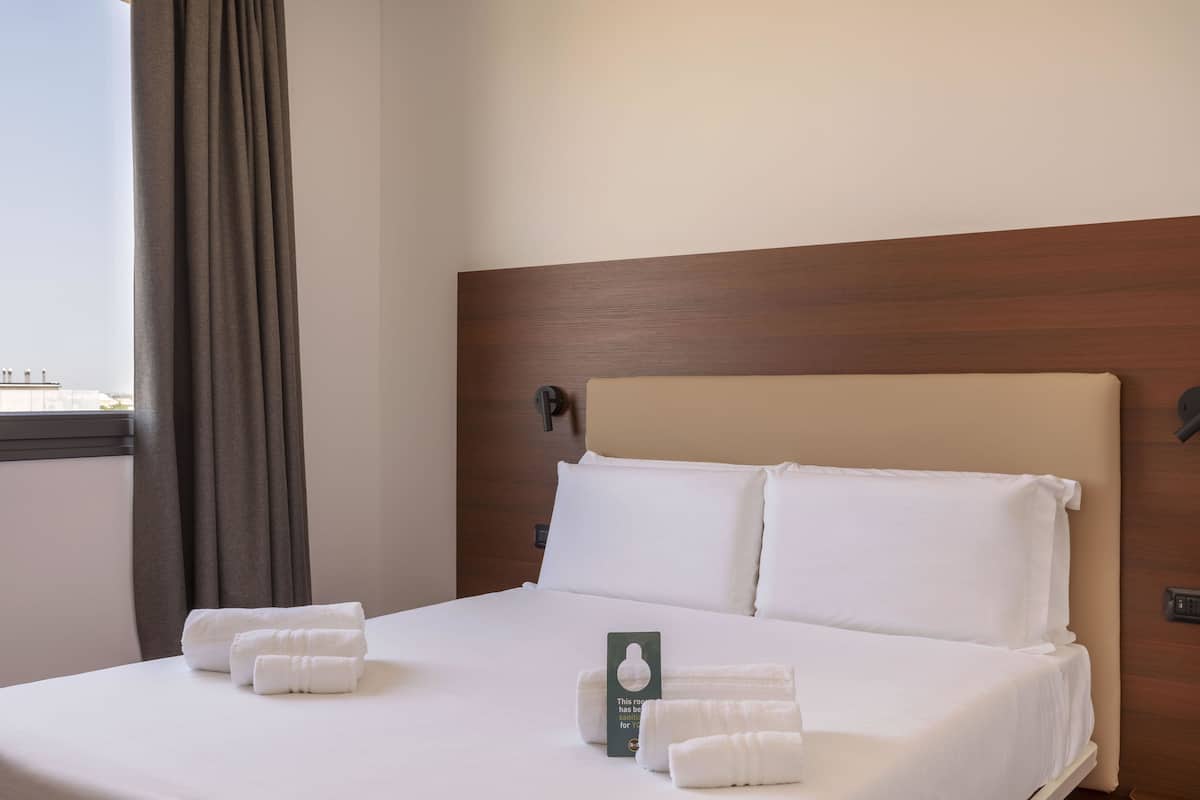 A neatly made bed with white linens and towels, a wooden headboard, and a view of the city through a window with curtains.